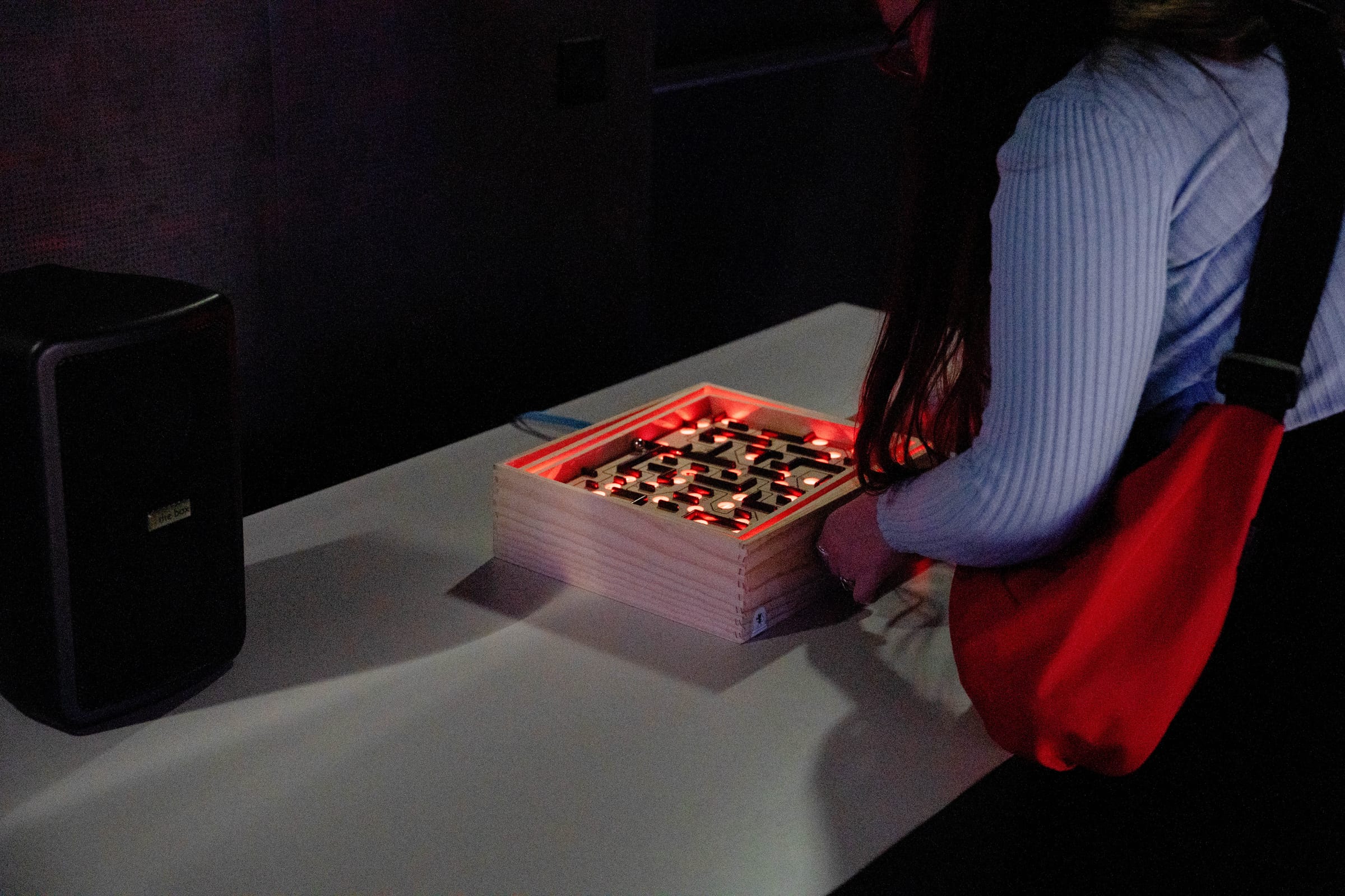 A player tilts the illuminated labyrinth board, its edge lit in red, connected by a cable to a small speaker on the table for the generative soundscape.