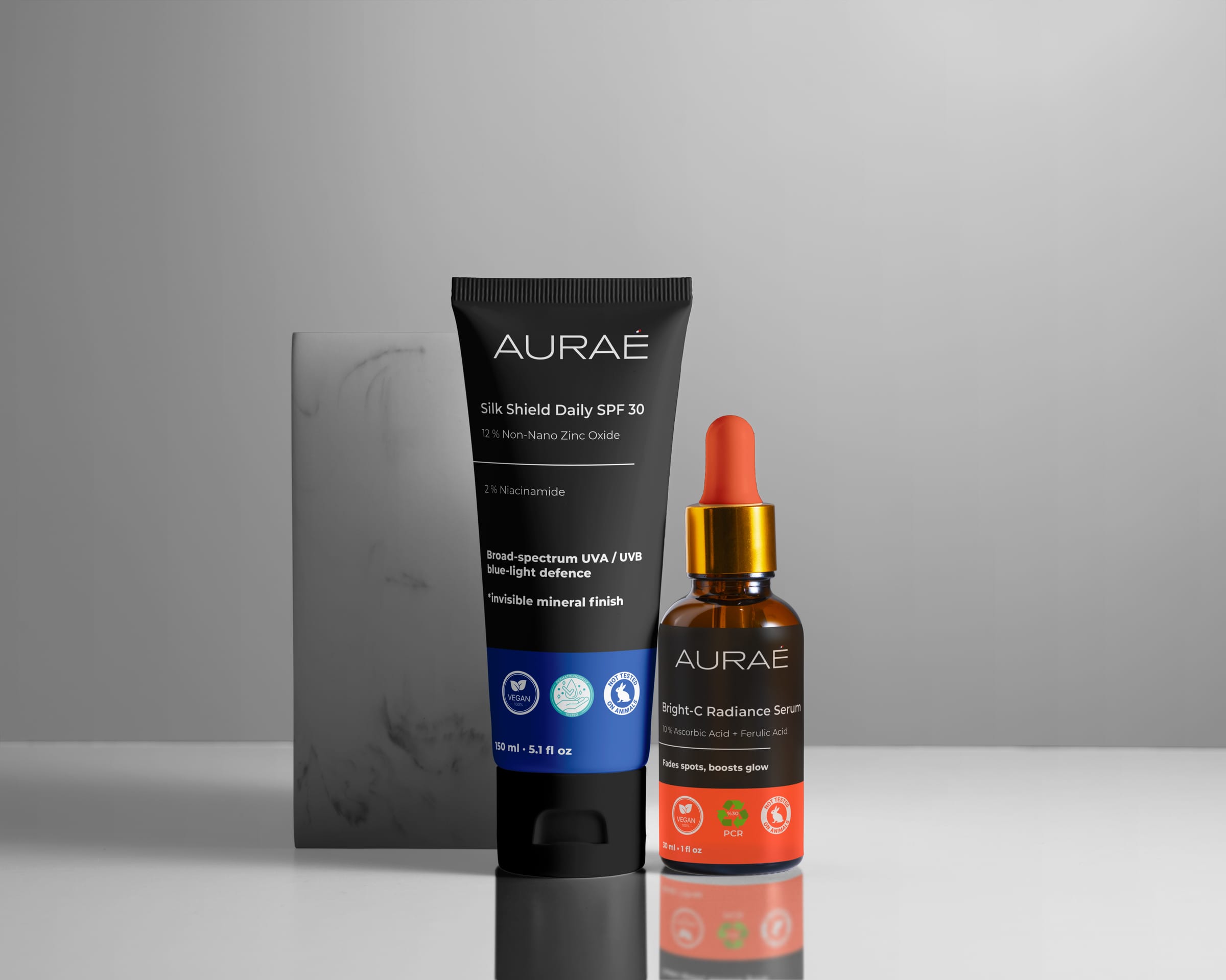 AURAÉ Silk Shield Daily SPF 30 tube beside Bright-C Radiance Serum, both using black labels with blue and coral accent bands.