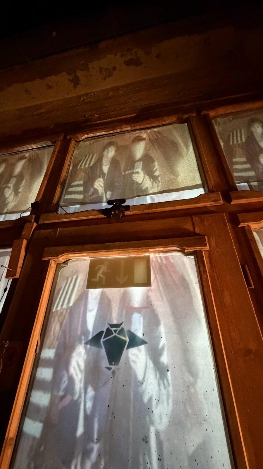 A close-up of one window pane showing the black owl-like emblem projected over a grainy human figure, an exit sign visible faintly through the glass.