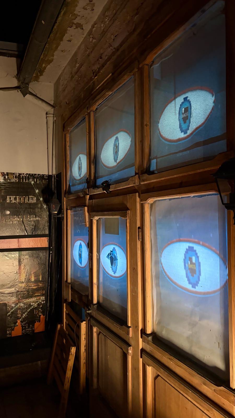A wide angled view of the six-pane window installation glowing blue with pixel-art eye motifs, in a dim room with other artwork on the walls.