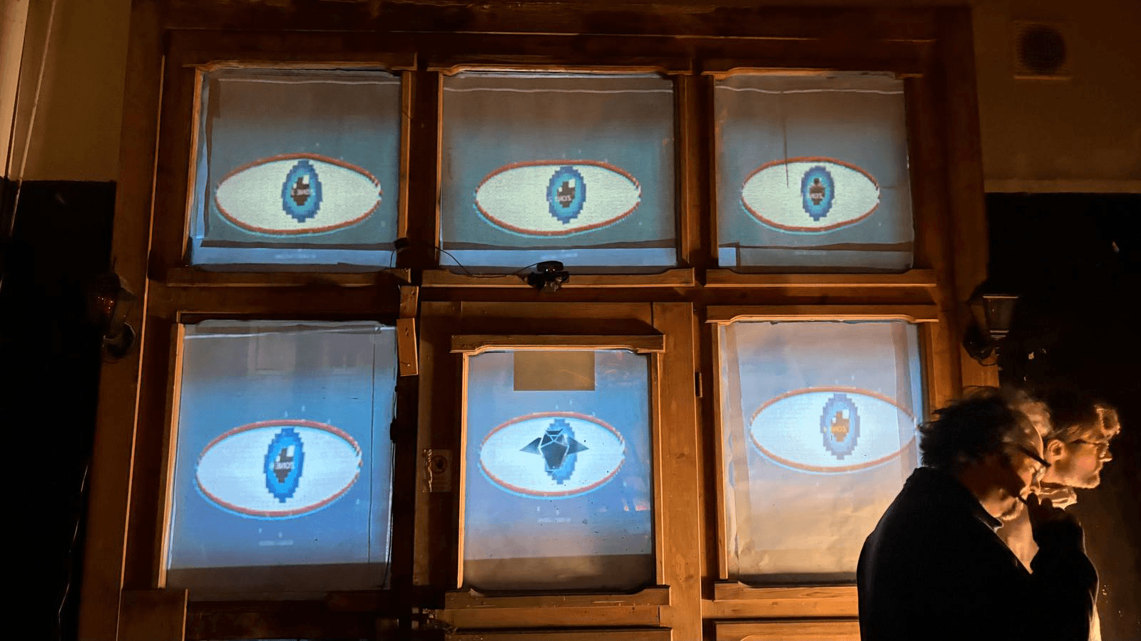 Six window panes used as a segmented display, each showing a large pixel-art eye motif in blue and red, with a black owl-like emblem projected in the centre pane.