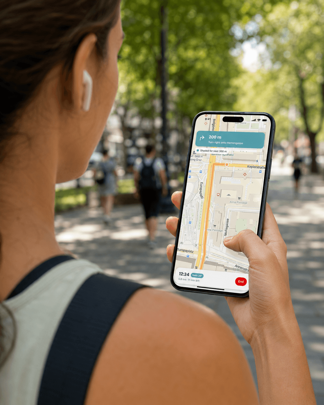 A woman checks turn-by-turn navigation on her phone while walking a tree-lined Graz street, the screen showing 'Turn right onto Herrengasse' and 'Shaded for next 300 m'.