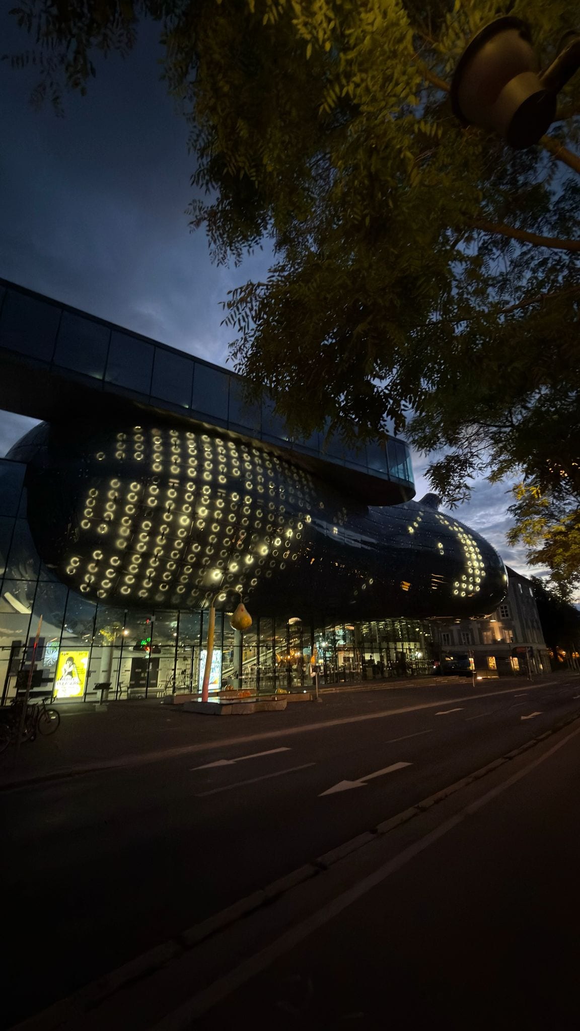 Street-level view of the Kunsthaus Graz building at dusk, the BIX facade glowing above the glass ground floor.